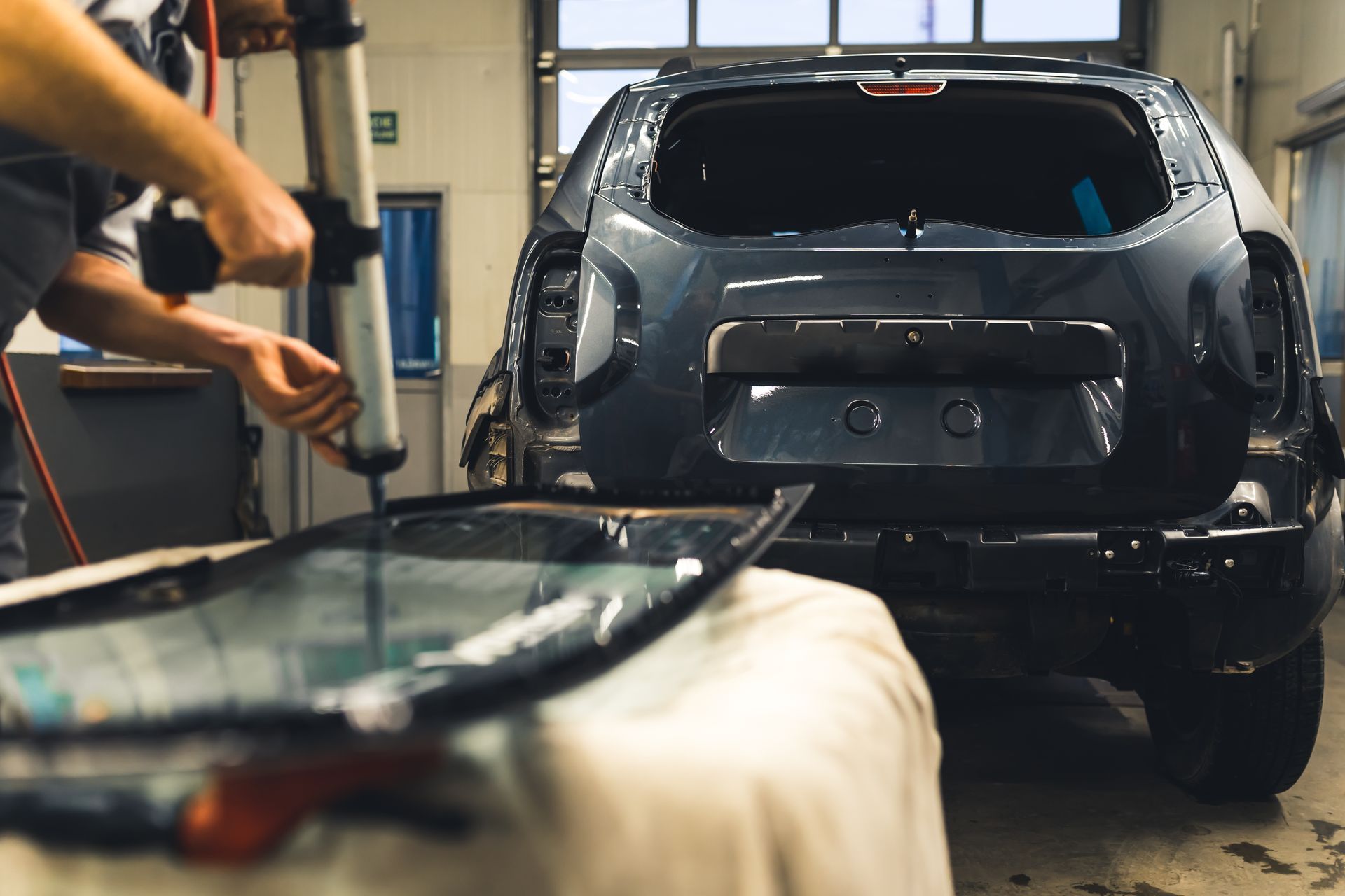 Une personne applique de l'adhésif sur une vitre de voiture, en préparation de son installation sur un SUV gris foncé dans un atelier de réparation.