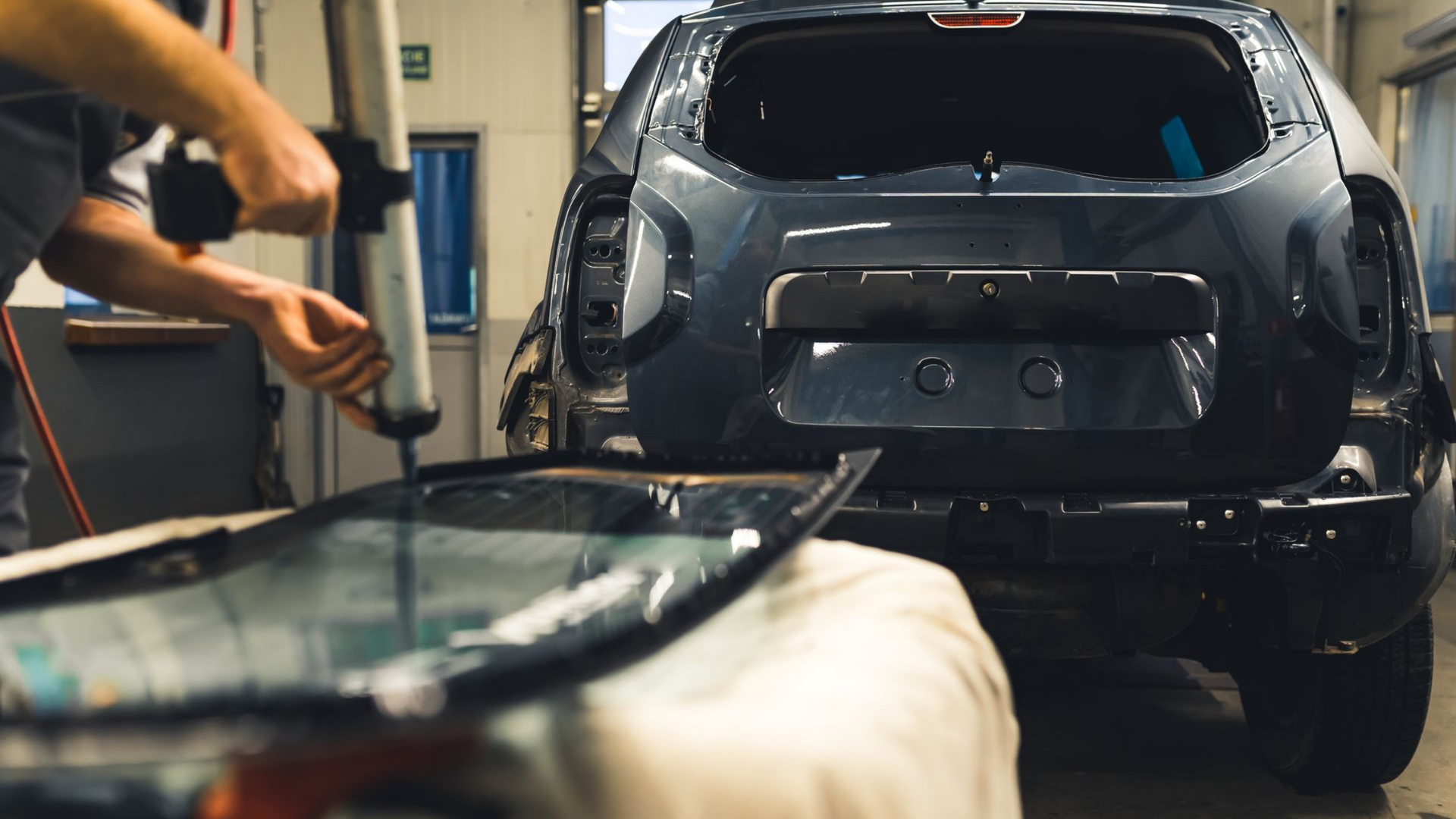 Dans un atelier, une personne applique du mastic sur la lunette arrière d'une voiture ; une autre voiture est visible en arrière-plan.