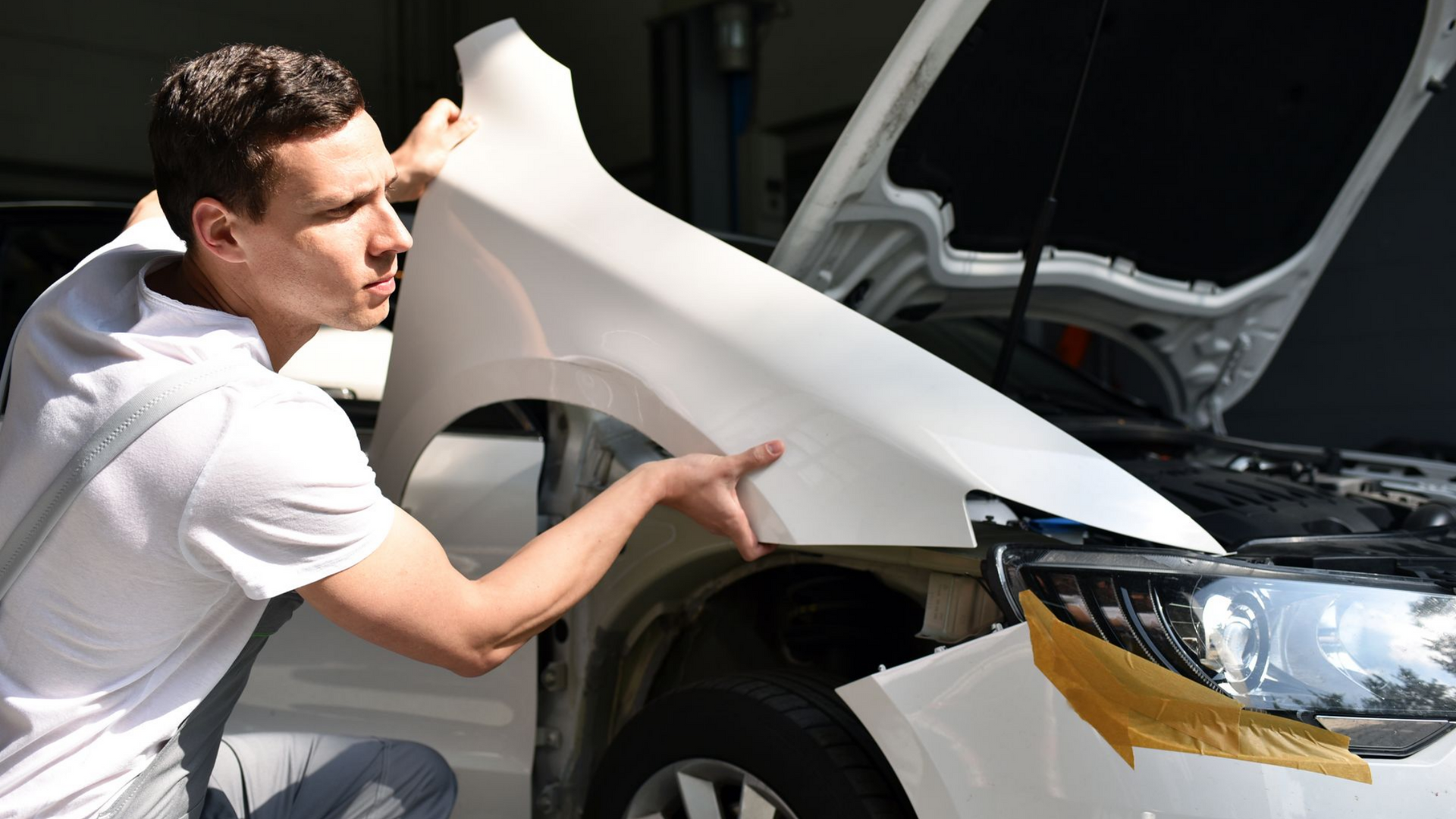 Un mécanicien installe une aile de voiture blanche dans un garage, capot ouvert.