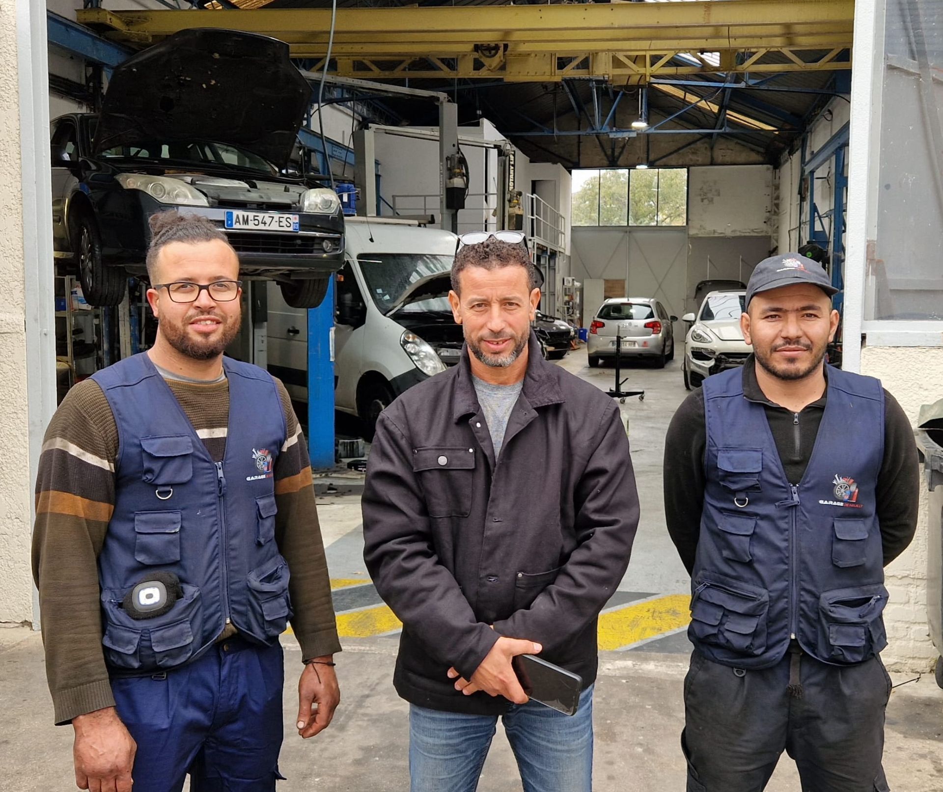 Un mécanicien sourit dans un garage, une voiture rouge derrière lui. Il porte une chemise rouge.