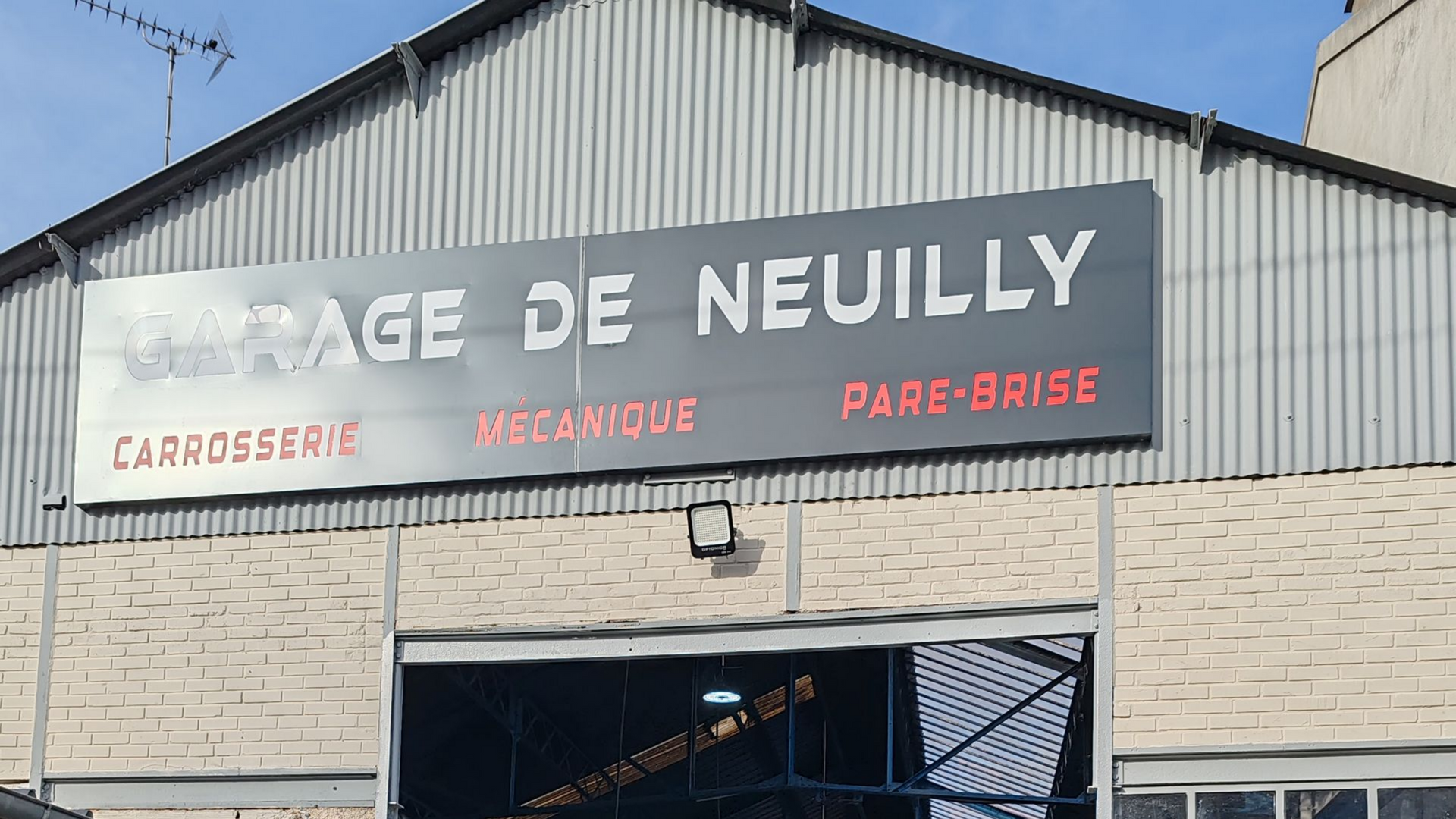 Panneau GARAGE DE NEUILLY au-dessus de la porte du garage, panneau gris avec texte blanc et rouge : Carrosserie, Mécanique, Pare-brise.