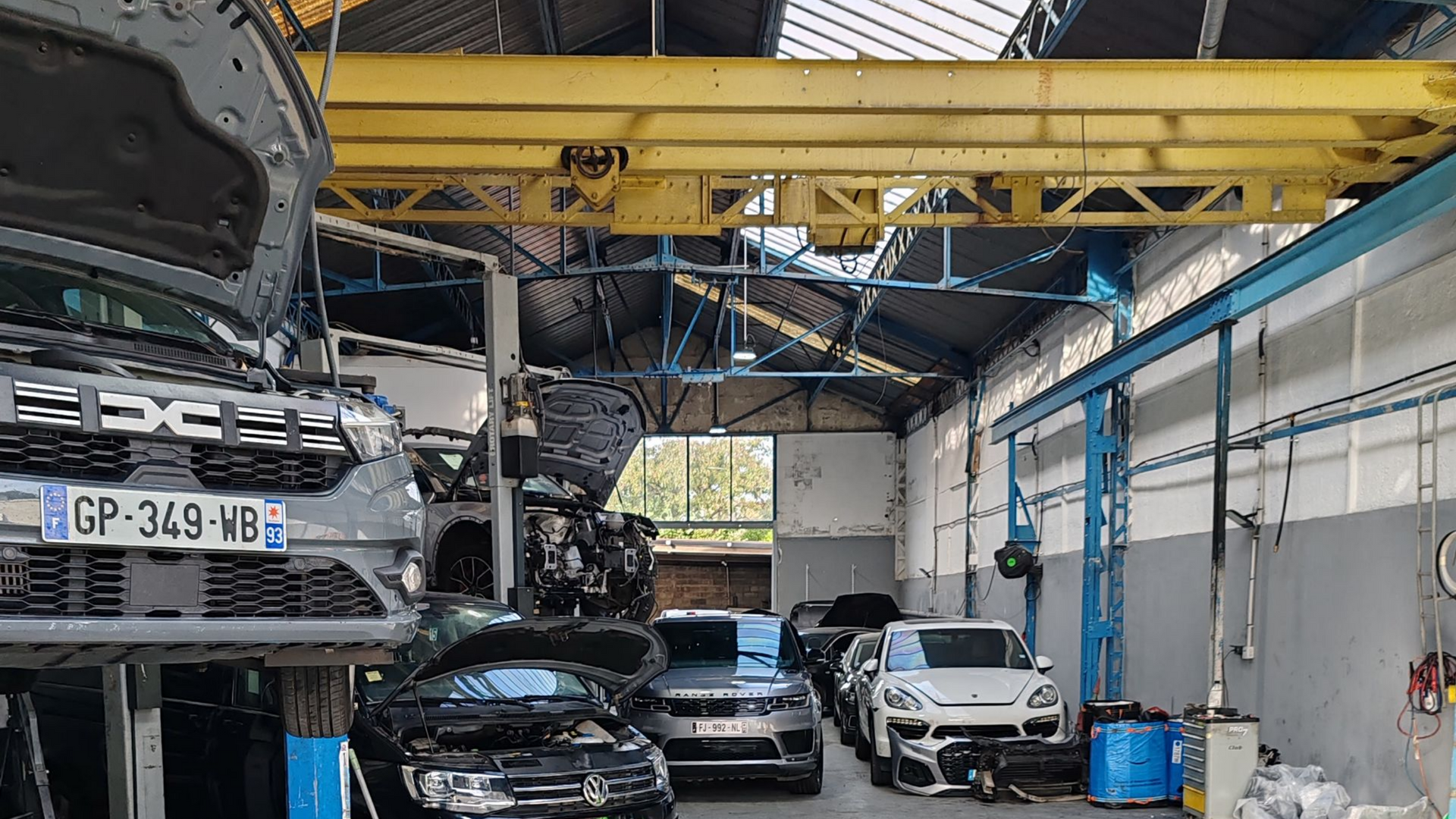 Des voitures à l'intérieur d'un garage, avec une grue, certaines sur des ponts élévateurs, d'autres garées.