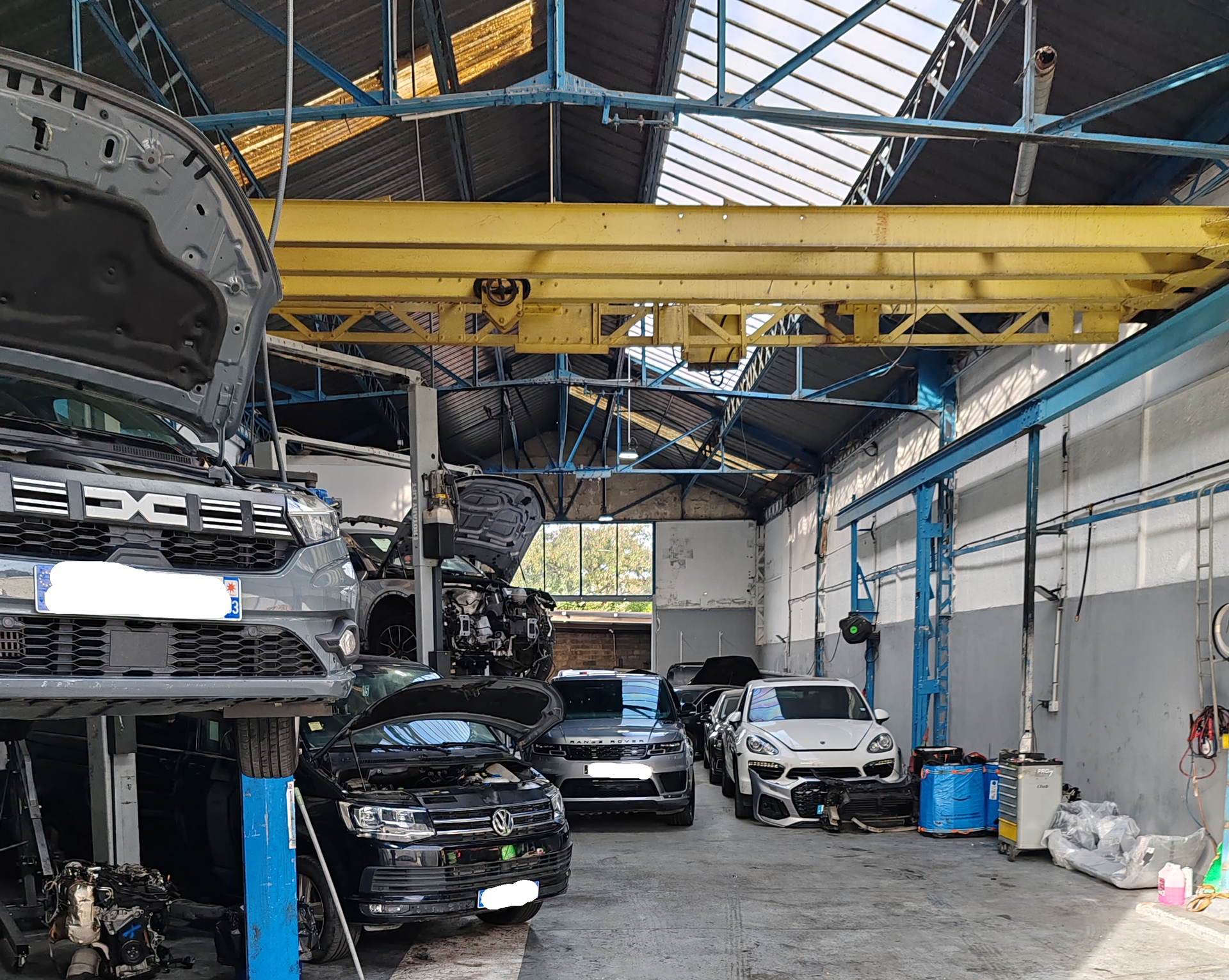 Intérieur d'un atelier de réparation automobile avec des véhicules sur des ponts élévateurs, un pont roulant et des voitures garées au sol.