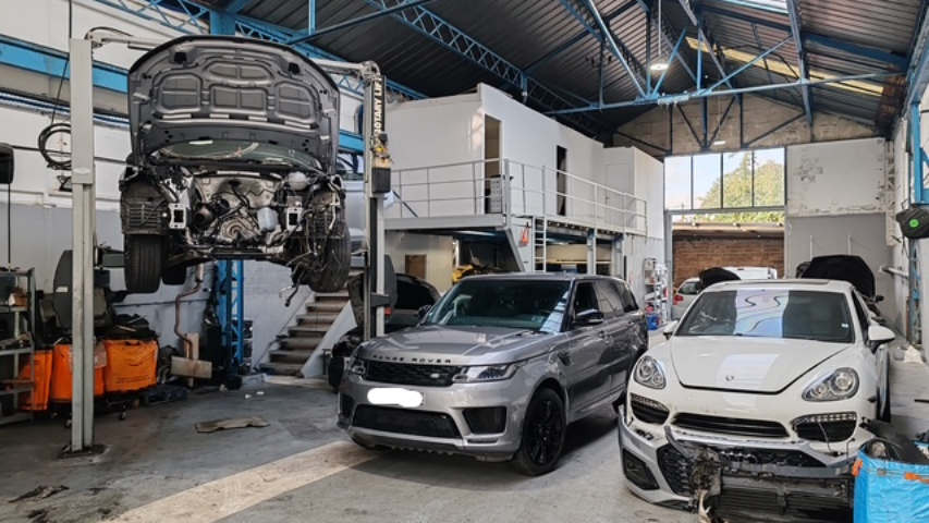 Atelier de réparation automobile avec une voiture sur un pont élévateur, une Range Rover et une Porsche Cayenne.