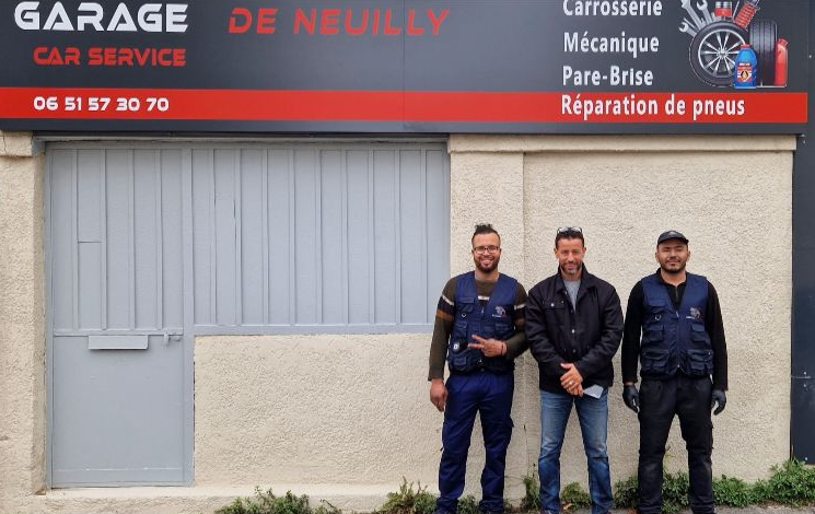 Un homme en tablier de jean sourit, les bras croisés, appuyé contre une camionnette blanche, à l'extérieur.
