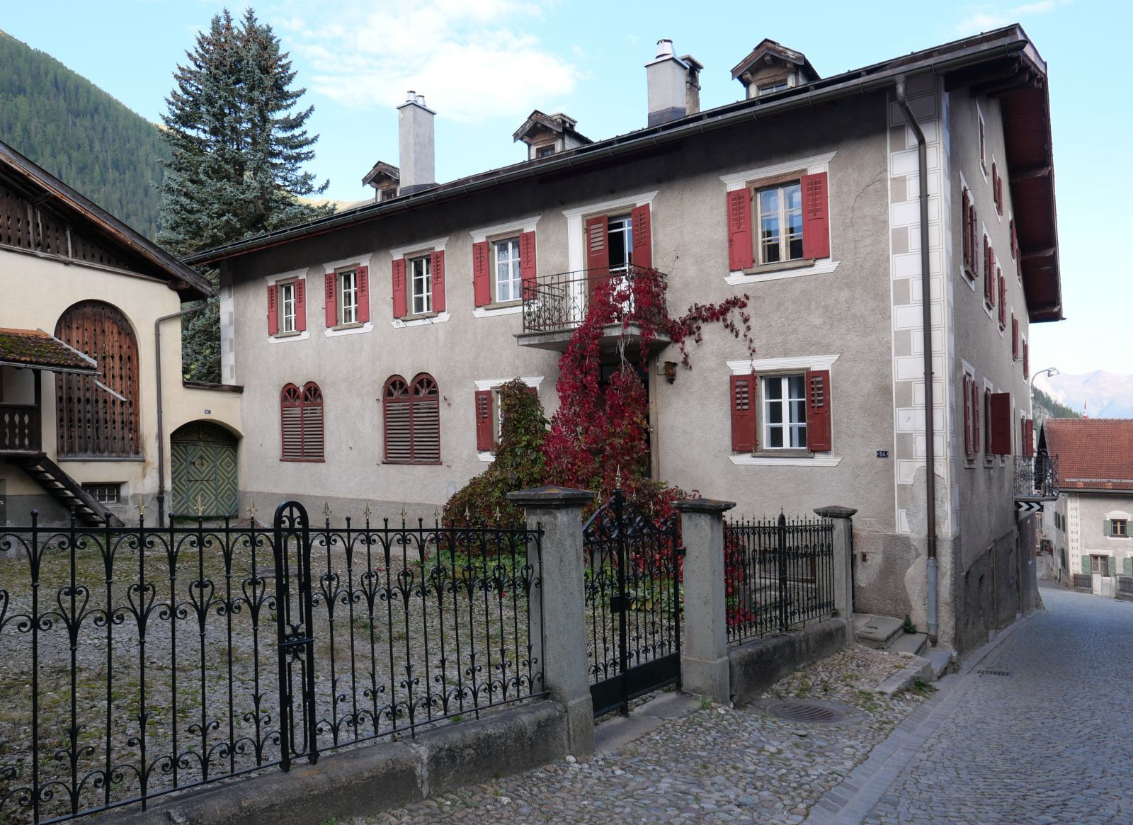 Ein grosses Haus mit roten Fensterläden, davor ein Hof mit Schmiedeisengitter