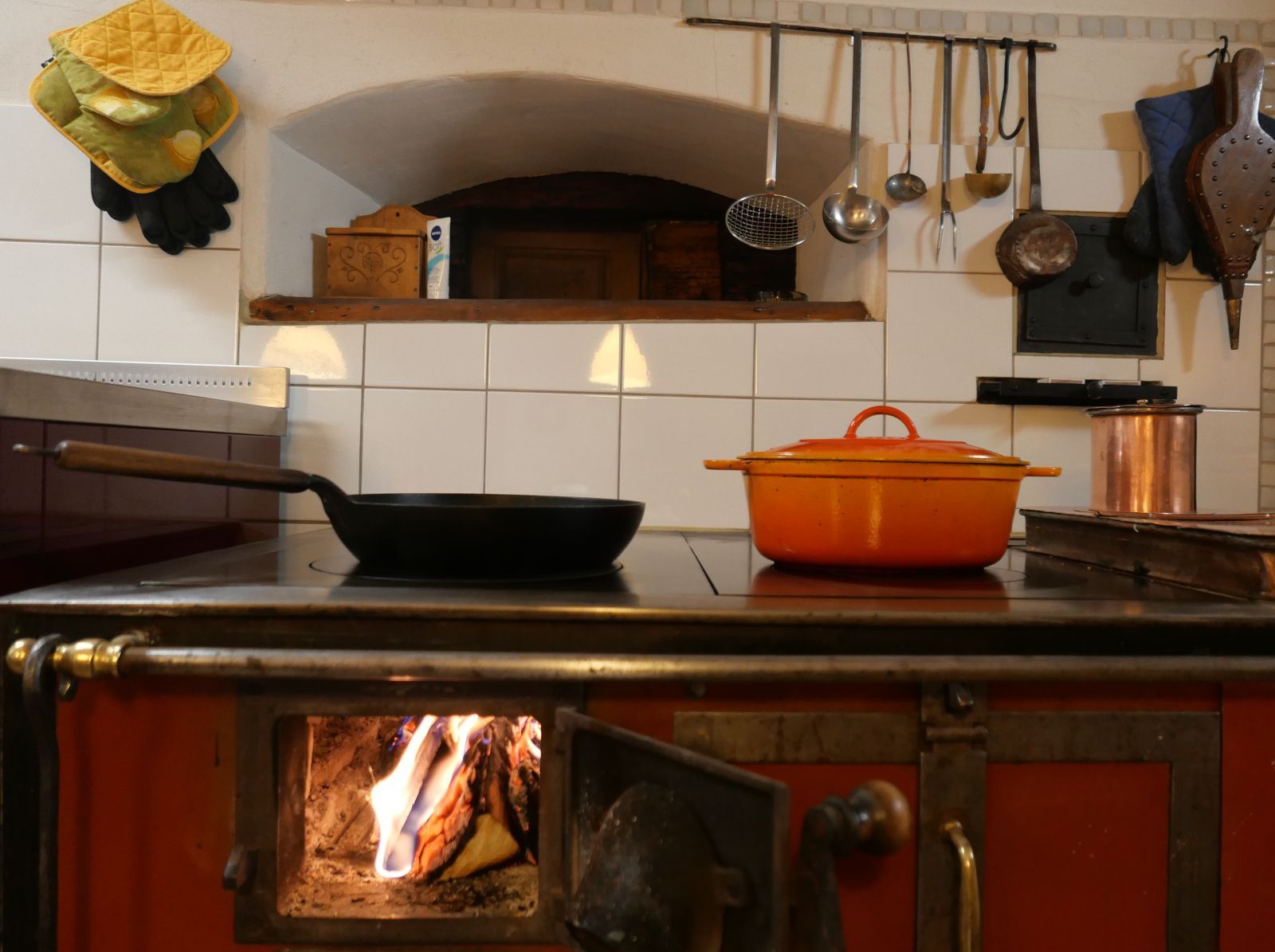 Ein Holzherd von vorne, im offenen Feuerloch brennt ein Feuer, auf der Kochplatte stehen eine Bratpfanne und ein Gusseisentopf.