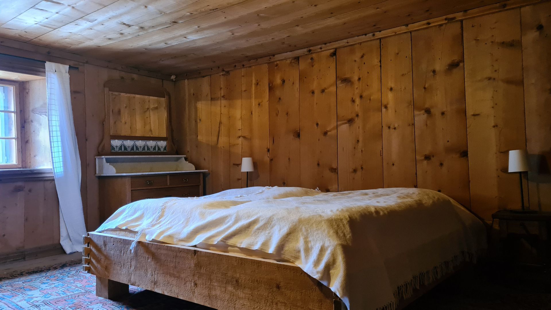 Ein Schlafzimmer mit Holzwänden und einem Doppelbett, in der Ecke eine Jugendstil-Waschkommode.