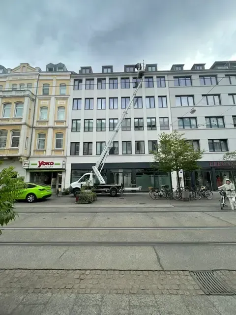 Ein LKW mit Hebebühne erreicht eine Gebäudefassade auf einer Stadtstraße. Bewölkter Himmel.