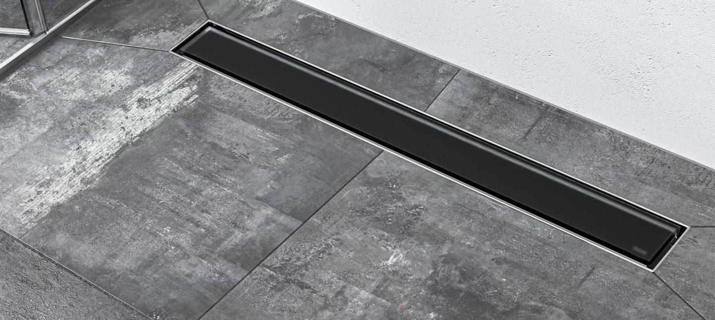 Desagüe oscuro y lineal en el suelo de la ducha alicatado. Los azulejos son grises y rectangulares, junto a una pared blanca.