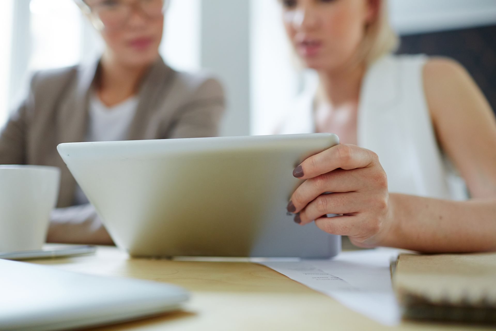 Dos mujeres sentadas a una mesa, mirando juntas una tableta, hablando de trabajo.