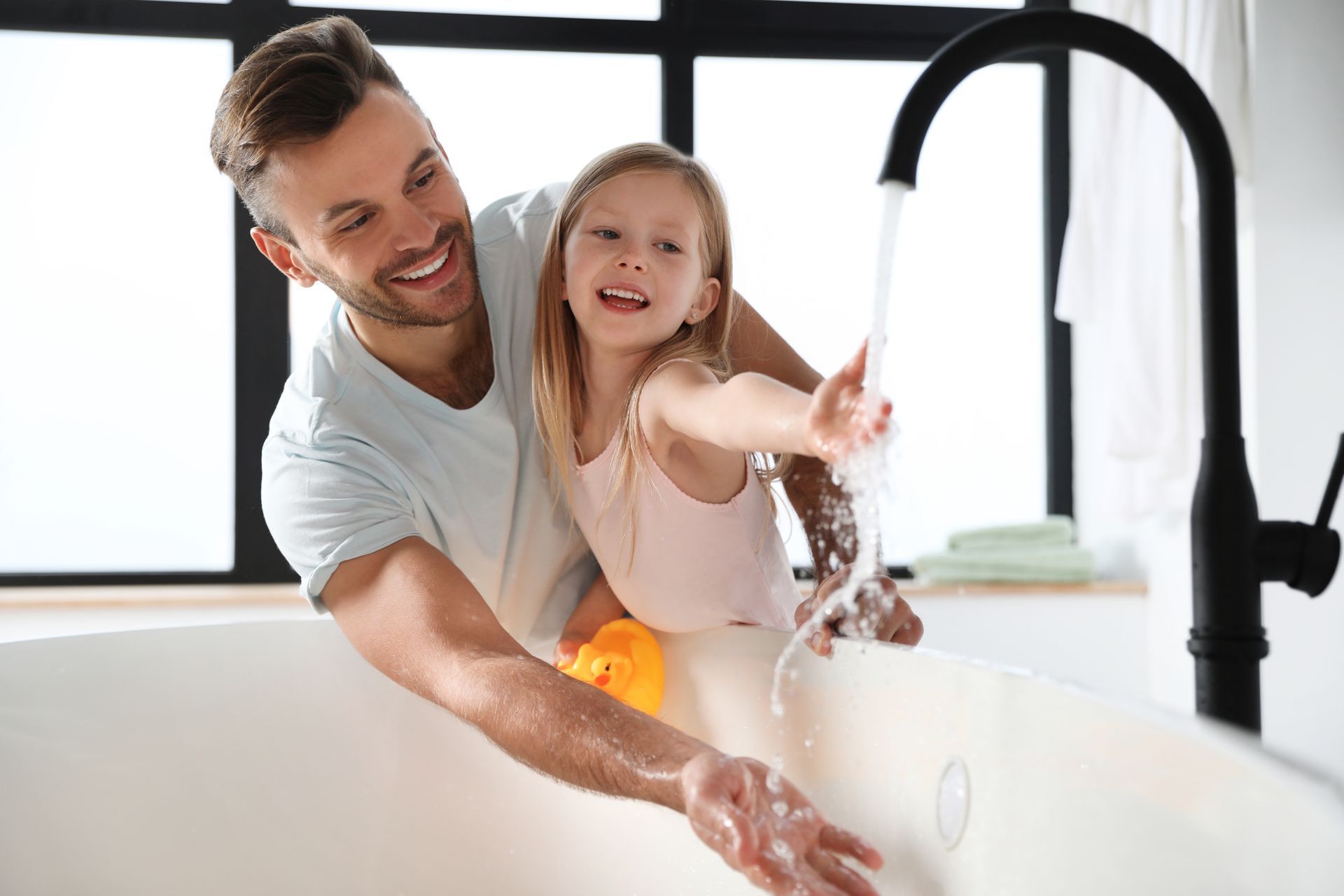 Un homme et un enfant jouent dans une baignoire où l'eau coule, souriant et tendant les bras.