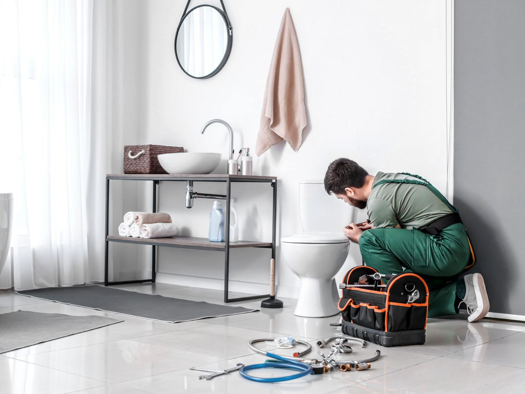 Un plombier en salopette verte travaille sur des toilettes dans une salle de bains lumineuse équipée d'une boîte à outils, d'un lavabo et de serviettes.