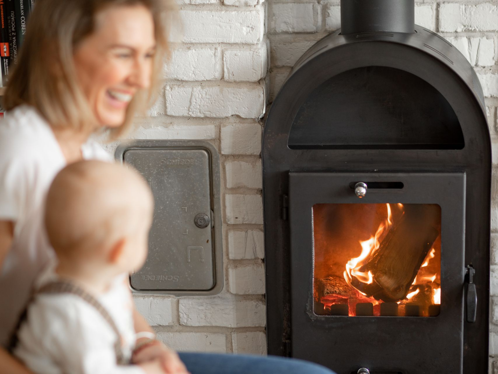 Une femme et son bébé sourient en regardant le feu de cheminée dans une pièce aux murs de briques.