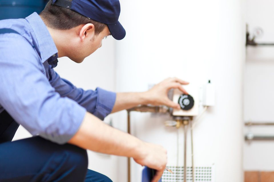 Un plombier en uniforme bleu ajuste le bouton de commande d'un chauffe-eau blanc dans une buanderie.