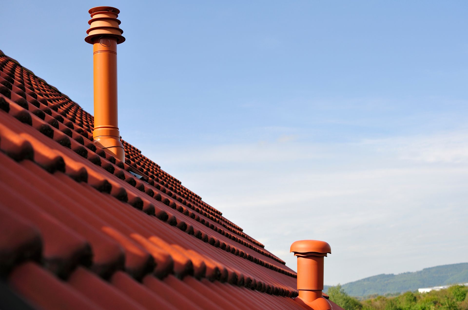 Toit de tuiles rouges avec deux cheminées orange sur fond de ciel bleu.