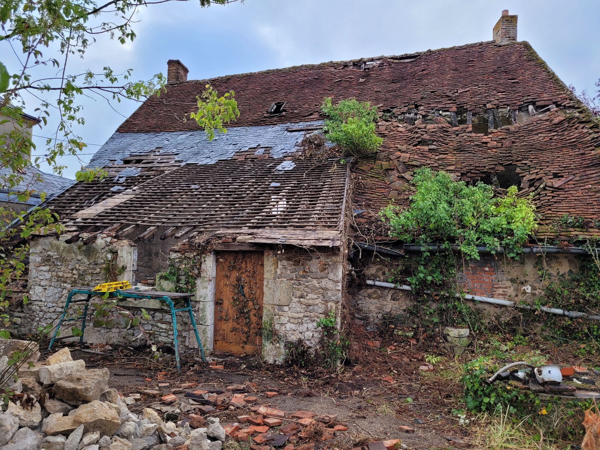 Maison en pierre délabrée, au toit effondré, envahie par la végétation, avec une porte en bois en façade.