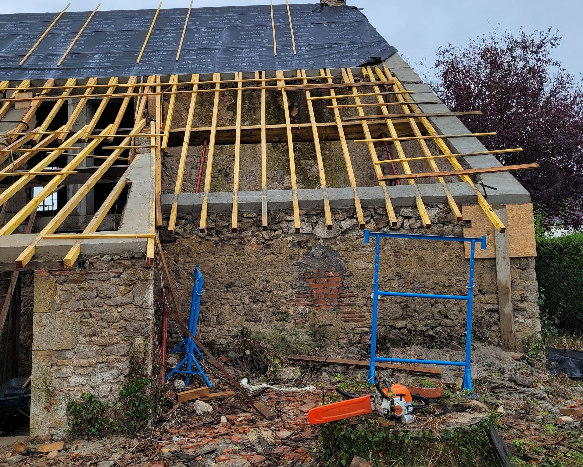 Toiture en pierre partiellement décapée d'une maison de campagne, avec chevrons jaunes et échafaudages bleus, dans une cour rurale