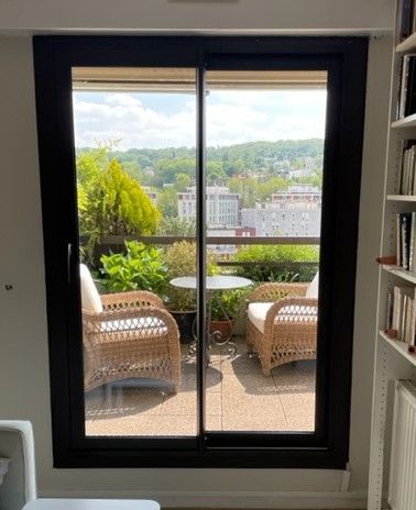 Fenêtre aux bords noirs donnant sur une terrasse avec une table et deux fauteuils d'extérieur
