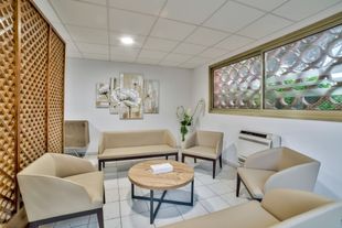 Salle d’attente lumineuse avec des fauteuils beiges, une table ronde, des œuvres murales et des fenêtres à motifs