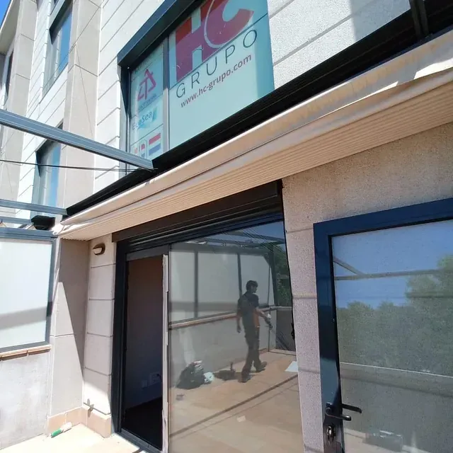 Exterior del edificio con toldo y persona reflejada en la puerta de cristal.
