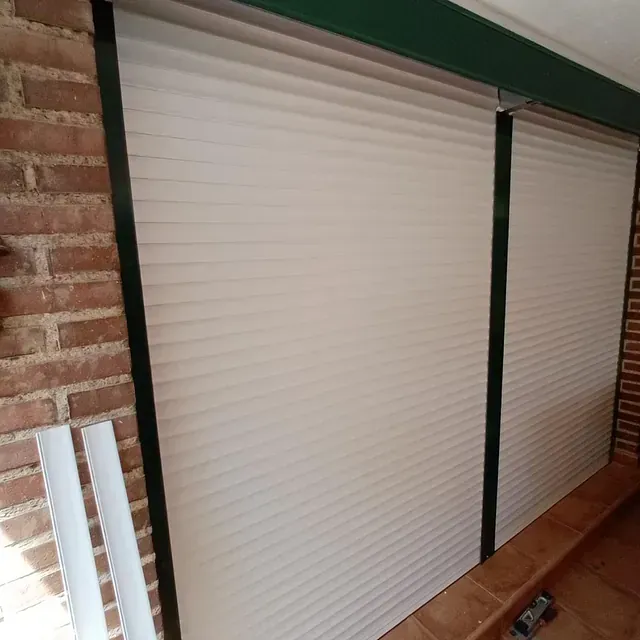 Persianas blancas plisadas que cubren una pared de ladrillo, sujetas a un marco verde.