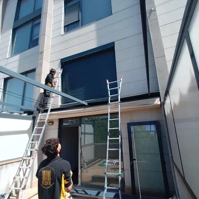 Obreros instalan una persiana enrollable de color oscuro en el exterior de un edificio, utilizando escaleras.