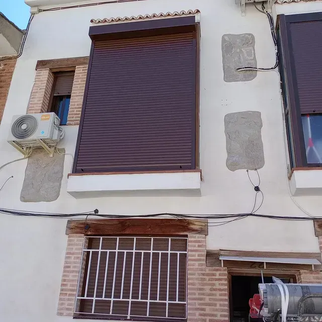 Exterior de un edificio con contraventanas marrones y unidad de aire acondicionado. Detalles de ladrillo y paredes blancas.