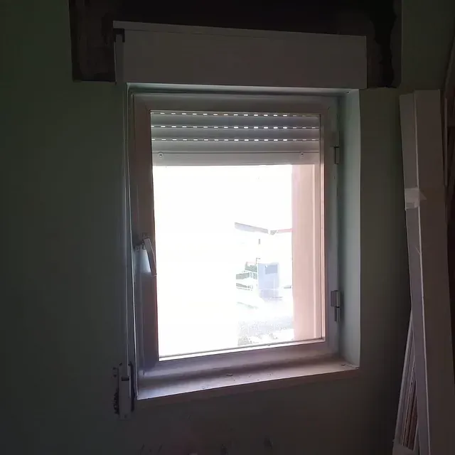 Ventana con estor enrollable gris claro parcialmente bajado, en una habitación verde claro.