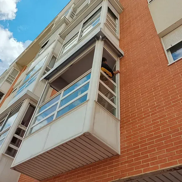 Persona limpiando una ventana en un balcón cerrado de un edificio de apartamentos de ladrillo.