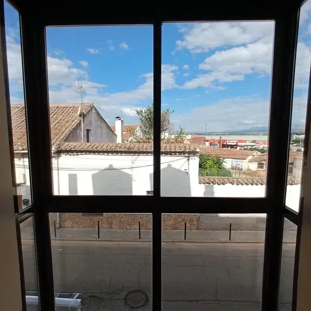 Vista a través de una ventana, mostrando tejados, calle y cielo azul con nubes.