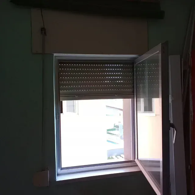 Ventana abierta con persiana enrollable cerrada. La luz entra a raudales desde el exterior.