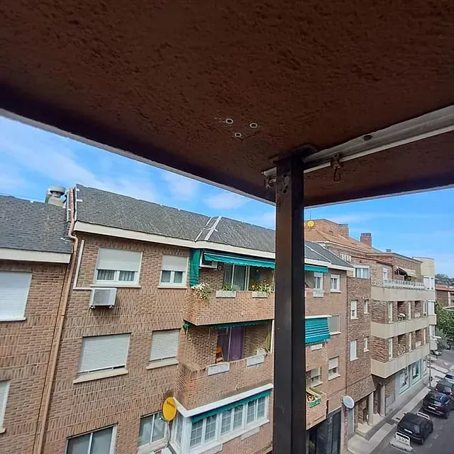 Vista de edificios de ladrillo y cielo azul desde un balcón con techo marrón y viga de soporte negra.