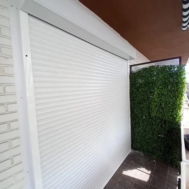 Persiana enrollable blanca en una pared de ladrillo, junto a un seto verde, bajo un toldo marrón.