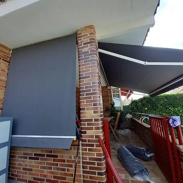 Un toldo gris oscuro da sombra a un porche de ladrillo. En primer plano se ve una barandilla roja.