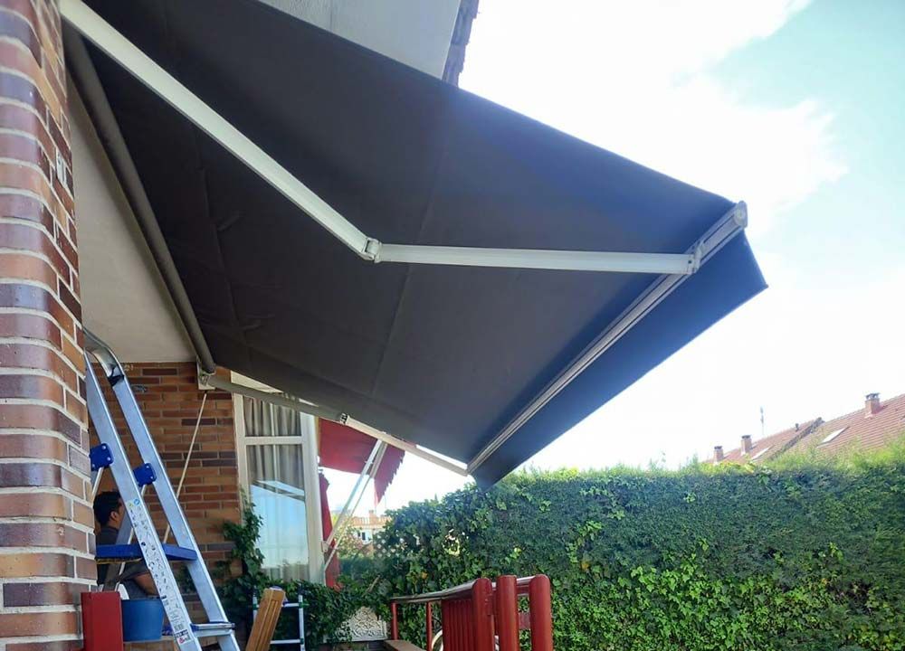 Un toldo retráctil marrón se extendía sobre una ventana, contra un cielo soleado y un seto verde.