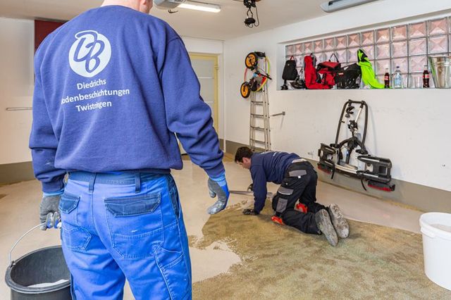 Zwei Arbeiter in einer Garage, einer kniet auf dem Boden, der andere steht daneben, Werkzeuge und Ausrüstung an der Wand.