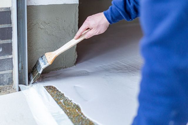 Eine Person verteilt Beton mit einer Kelle auf einem Boden neben einer Wand.