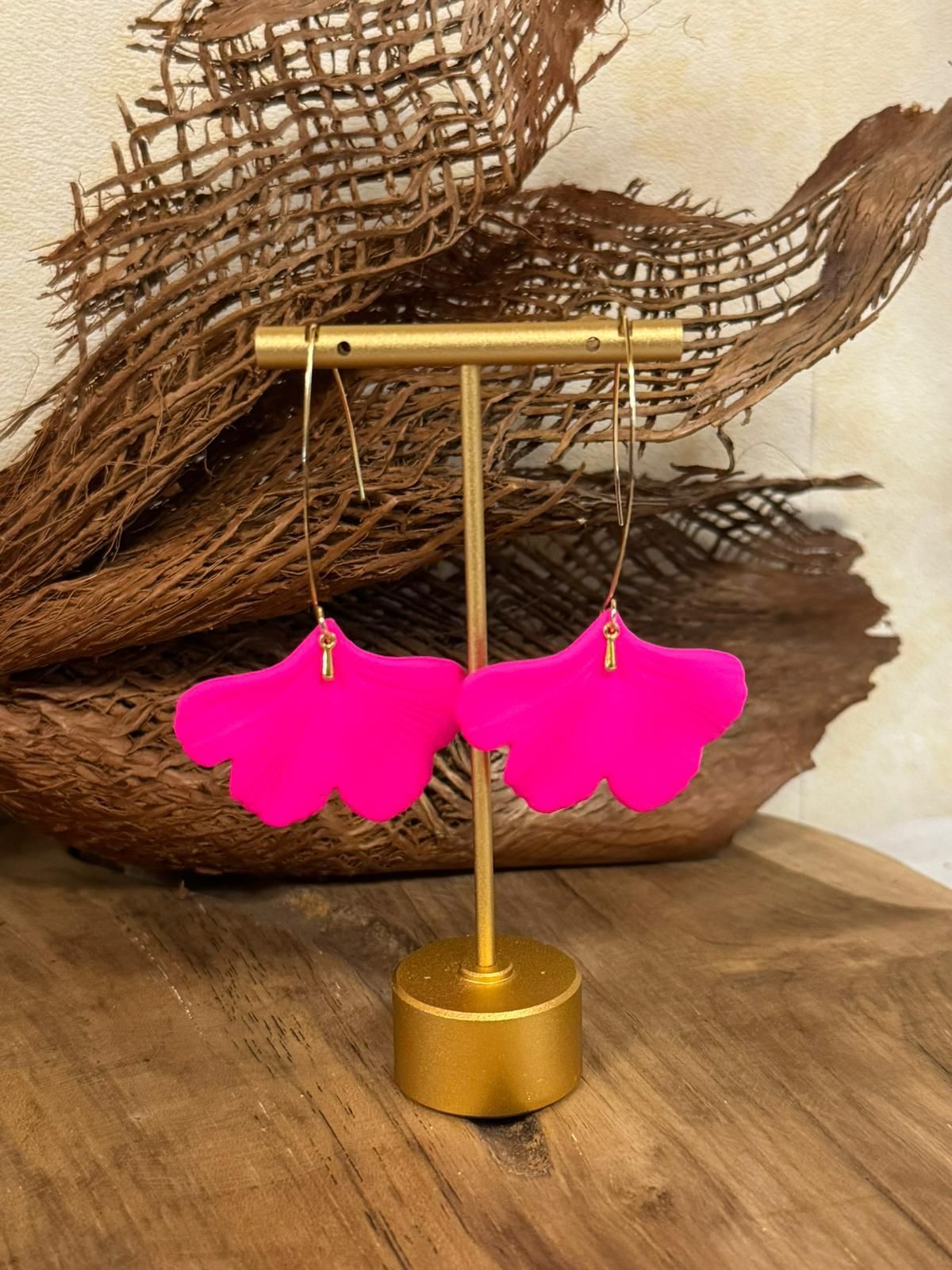 Boucles d'oreilles en forme de feuille rose suspendues à un support en or, sur un fond brun flou.