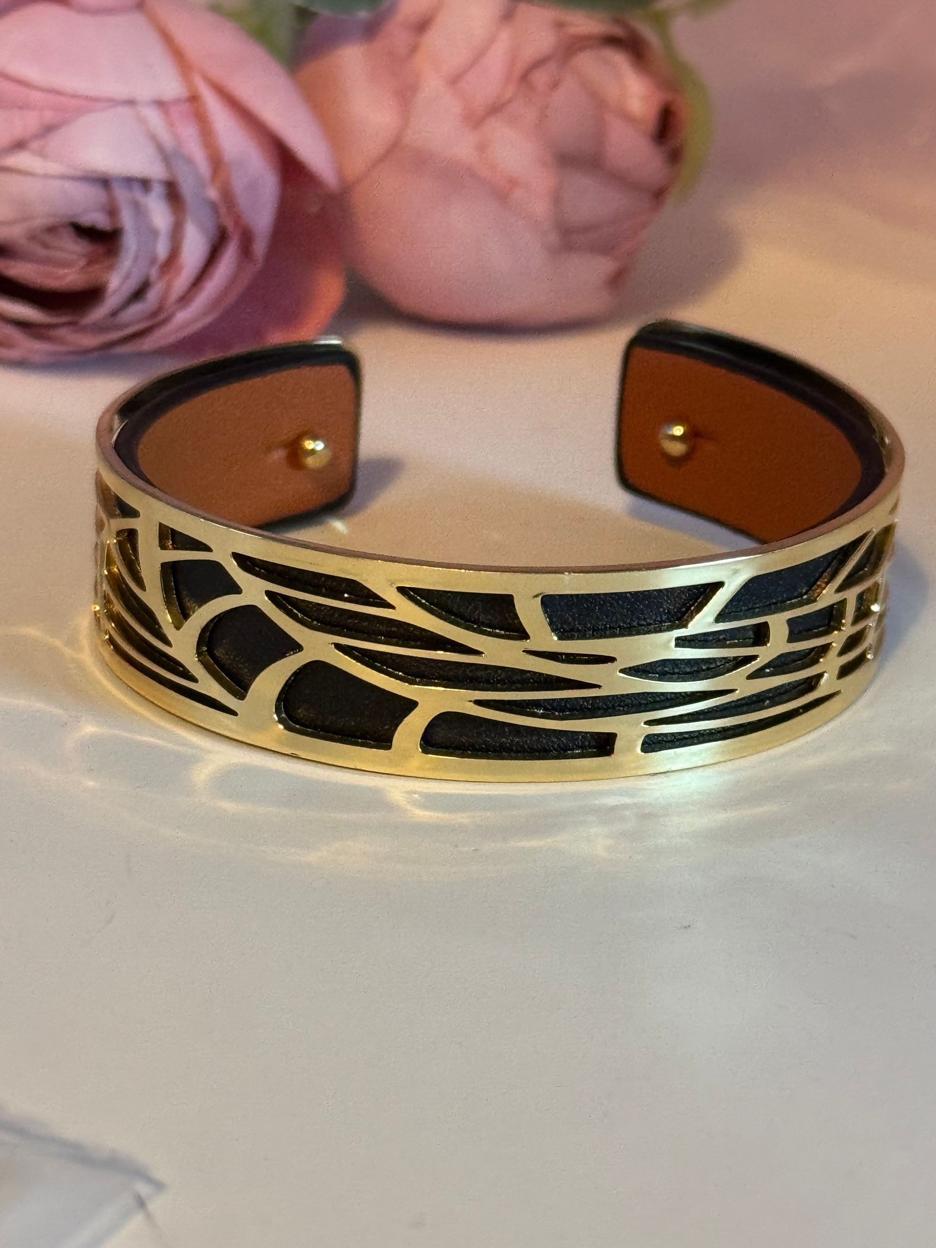 Bracelet manchette or et noir à motif ajouré, sur fond blanc avec des fleurs roses en arrière-plan.