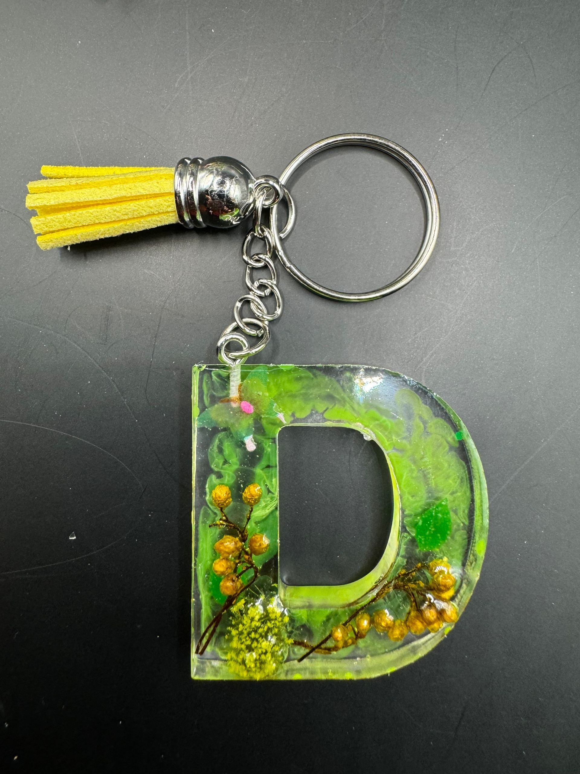Porte-clés en forme de D, rempli de résine verte et de fleurs séchées, attaché à un pompon jaune et une chaînette argentée.