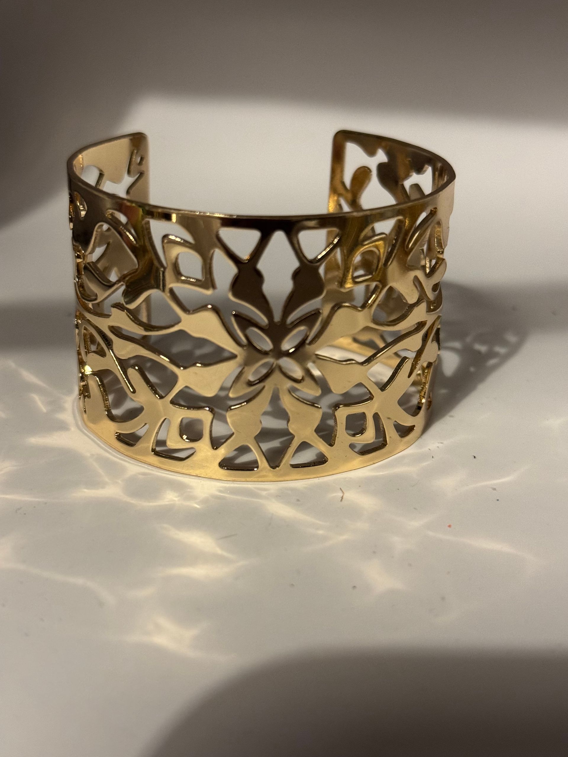 Bracelet manchette doré à motif floral ajouré, posé sur une surface claire et projetant une ombre.
