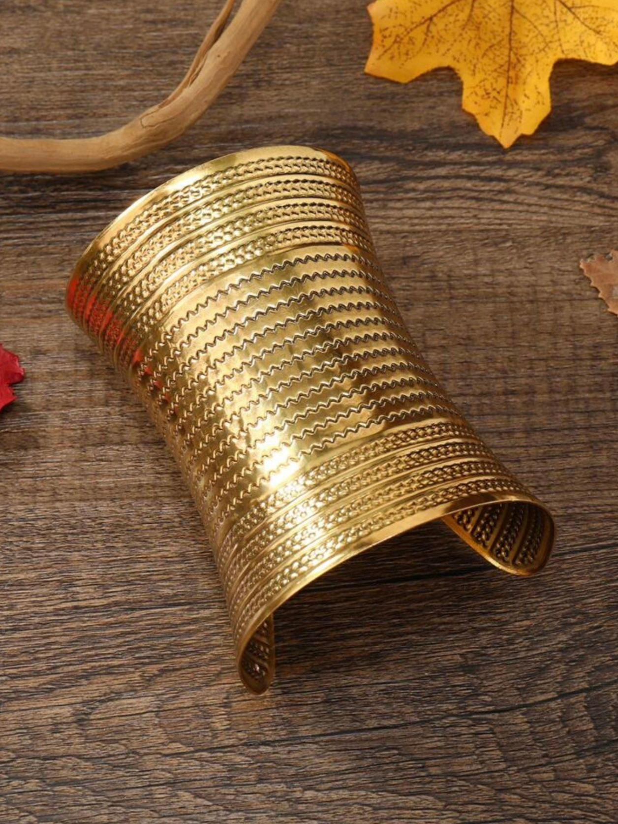 Bracelet manchette doré à surface texturée, posé sur un support en bois orné de feuilles.