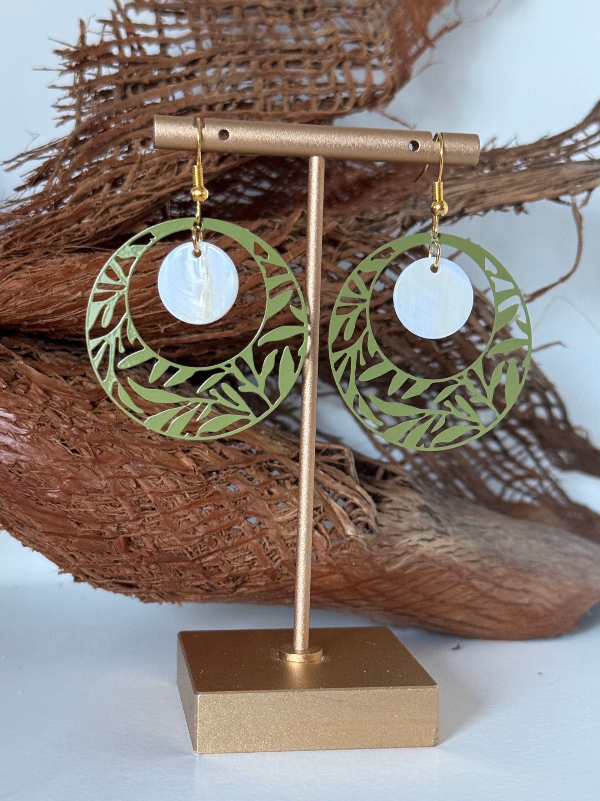 Paire de boucles d'oreilles en forme de feuille verte avec un centre circulaire blanc, présentées sur un support doré.