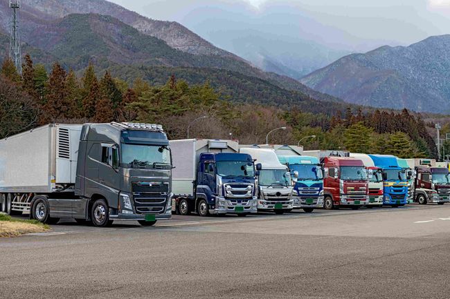 Una fila de camiones semirremolques estacionados en un estacionamiento con montañas al fondo.