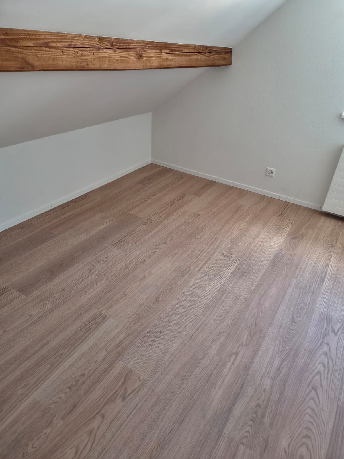 Leeres Dachzimmer mit schrägen weissen Wänden, freiliegendem Holzbalken und hellem Holzboden