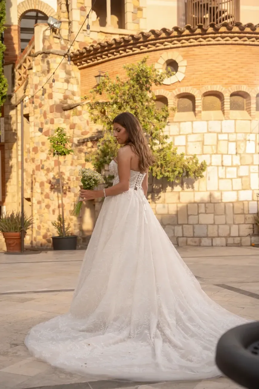 Mujer con vestido de novia blanco sosteniendo flores, de pie en un patio.