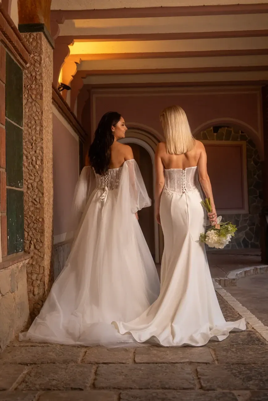 Dos mujeres con vestidos de novia caminan por un pasillo arqueado
