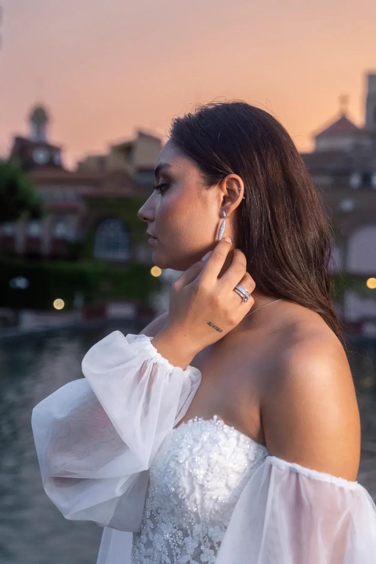 Mujer con vestido blanco se ajusta un pendiente, posando al aire libre. Al atardecer y con edificios de fondo.