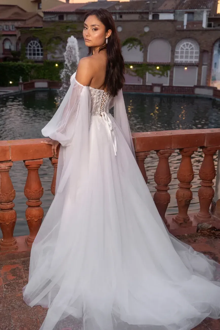 Mujer con un vestido de novia blanco con hombros descubiertos, mirando por encima del hombro a un balcón junto a una fuente.